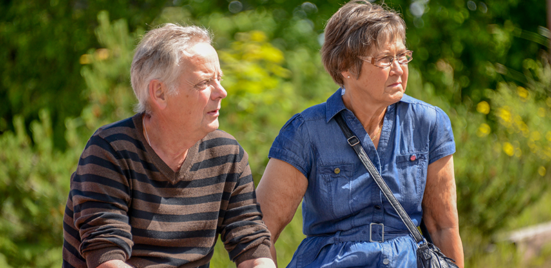 Ett äldre par sitter utomhus en solig dag. Mannen bär en brun- och svart-randig tröja, och kvinnan har en blå jeansklänning och glasögon. De tittar båda åt samma håll. I bakgrunden finns grönskande träd.